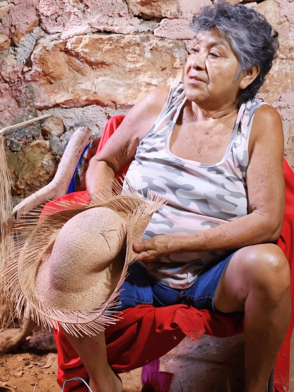 Eine mexikanische Frau mit schwarzen, graumelierten Haaren, sitzt in einer Kellerhöhle auf einem Stuhl und zeigt einen naturfarbenen Strohhut.